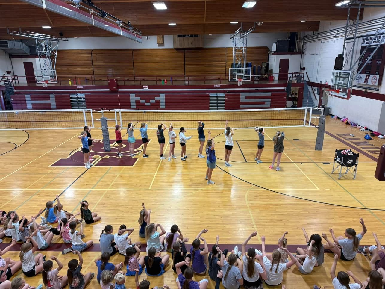 Dozens of young players cheering from the floor during a volleyball camp in the Hamilton Middle School gym